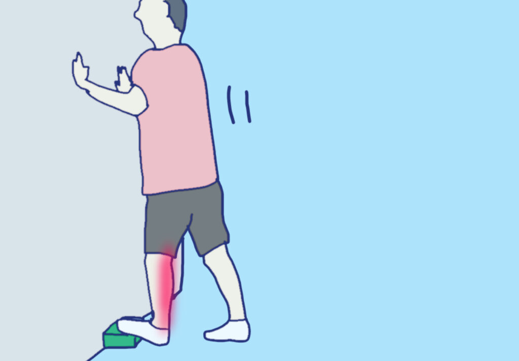 壁際にヨガブロックを置いて、ブロックに左足のつま先をかけ、骨盤を壁に近づけることで下腿三頭筋をストレッチしている